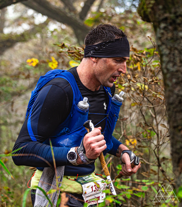 Trail runner in a blue hydration vest carrying poles on a wooded mountain course